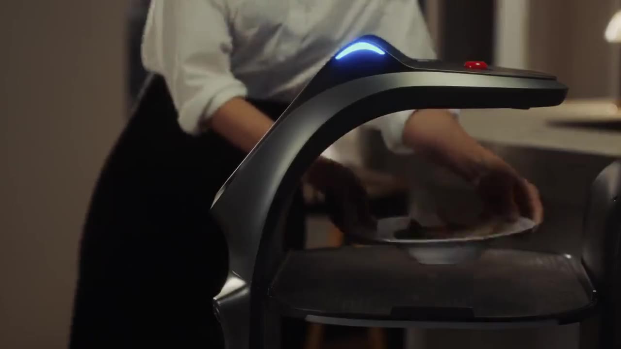 Manufacturer footage from Pudu: a server sets a plate on BellaBot Pro's tray at the kitchen pass